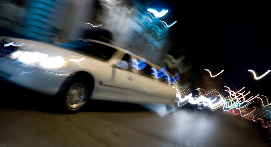 White limousine driving through a city at night, a blur of lights visible in the background.