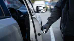 A man's gloved hand wearing black clothes opening a silver car door depiction of car getting stolen