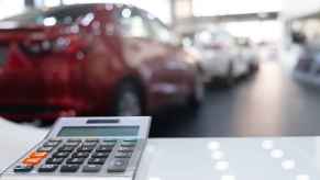 A blurred car dealership showroom with a desk calculator in the foreground to depict a car payment