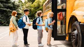 Row of students wait to board a bright yellow school bus, trees visible in the background.