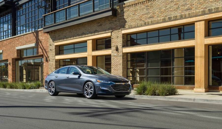 A gray Chevrolet Malibu parked alongside a large building.