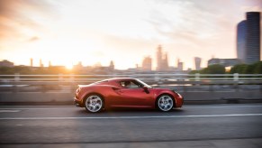 Red Italian Alfa Romeo sportscar driving across a bridge, a city skyline visible in the background.