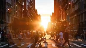 Pedestrians jaywalking across a New York City street, the sun setting in the background.