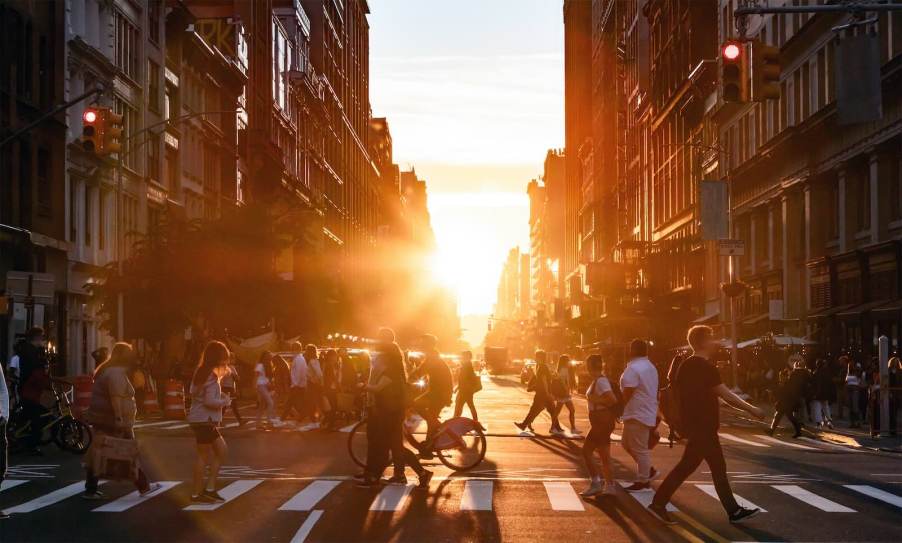 Pedestrians jaywalking across a New York City street, the sun setting in the background.