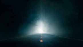 A foggy highway at night with light shining at the viewer down the road depiction of the "black dog" urban legend in trucking