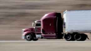 A semi-truck with dark purple cab and white trailer driving fast on highway in left profile view