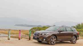 A dark brown Subaru Outback midsize SUV parked in left profile view at a Pacific Northwest-looking beach