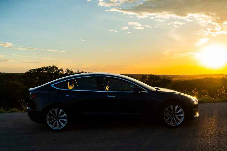 A dark-colored Tesla EV parked in full right profile view at sunset