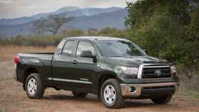 A dark-colored 2013 Toyota Tundra parked in right front angle view on sandy ground with green trees in background