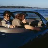 James Bond and his passenger look backward out of a BMW Z3 convertible during the "Goldeneye" film, the ocean visible in the background.