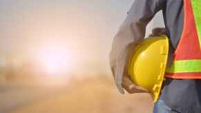 A road worker shown from shoulders down holding a hard hat against his left hip