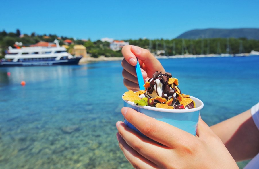 The hands of a woman eating ice cream out of a bowl, a cruise ship parked in a bay in the background.