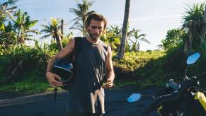 Long-haired man stands next to a motorcycle while holding his helmet.