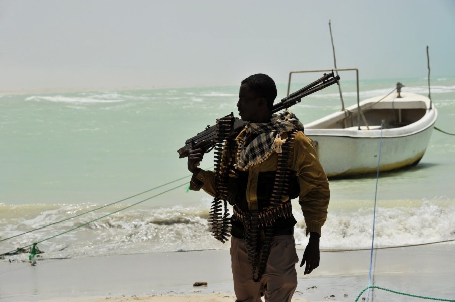 Somali pirate holding a machine gun on the beach, his boat anchored in the background.