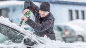 A man clears snow off a car windshield while making an exaggerated, wildly grimaced face