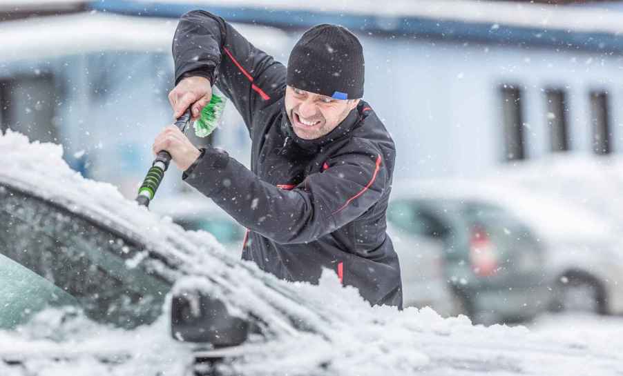 A man clears snow off a car windshield while making an exaggerated, wildly grimaced face