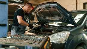 An automotive technician leaning over a vehicle using a wrench, with a cart full of tools at the ready