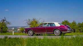 A 1984 Cadillac Seville driving down a scenic road as part of a car show