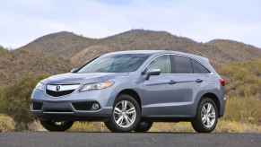 A light silvery-blue 2015 Acura RDX parked in left front angle view in front of grassy green mountains