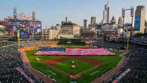 A huge USA national flag unfolded at baseball stadium, the Detroit city skyline visible in the background.