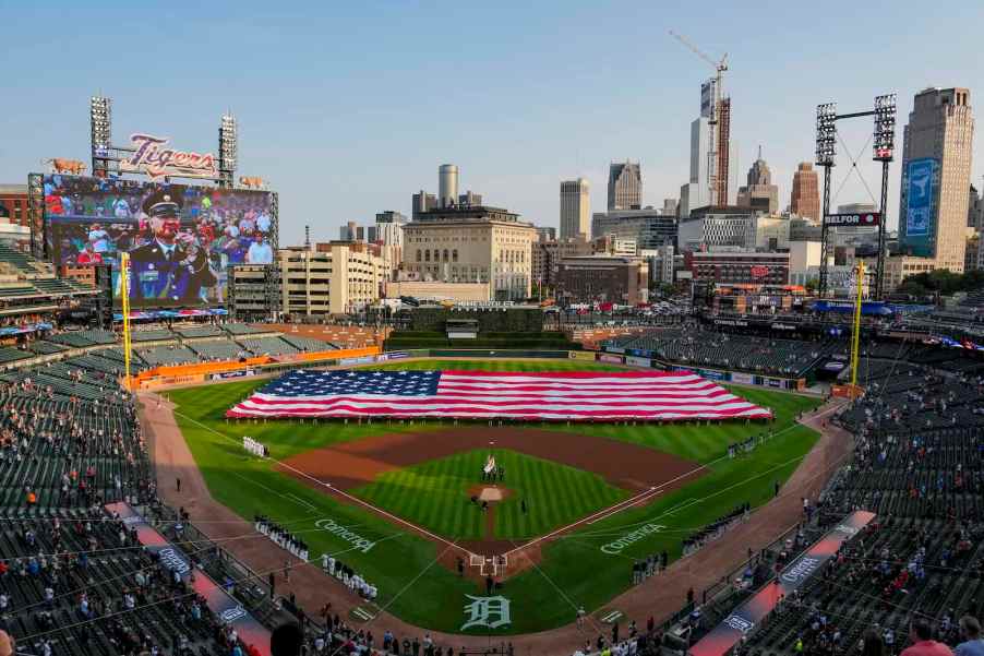 A huge USA national flag unfolded at baseball stadium, the Detroit city skyline visible in the background.