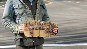 Homeless man standing on the street with an "anything helps" cardboard sign, cars in the background.