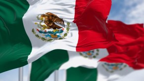 Row of Mexican national flags blowing in the wind, a blue sky visible in the background.