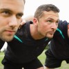 Line of rugby players link arms in a scrum formation, grass visible in the background.