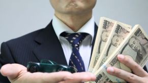 A man holding up a toy car and stacks of money, representing a scam involving cars