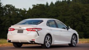 A whit 2018 Toyota Camry is parked in front of pine trees in right rear corner view