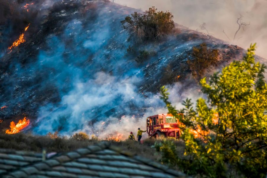 Two firefighters stand next to the LA County "Fire Dozer" while a hillside burns in the background.