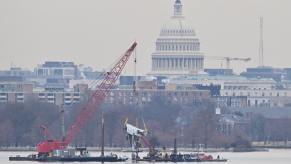 Crane on a barge lifts the wreckage of a plane crash out of the Potomac River, the Capital Building in the background.