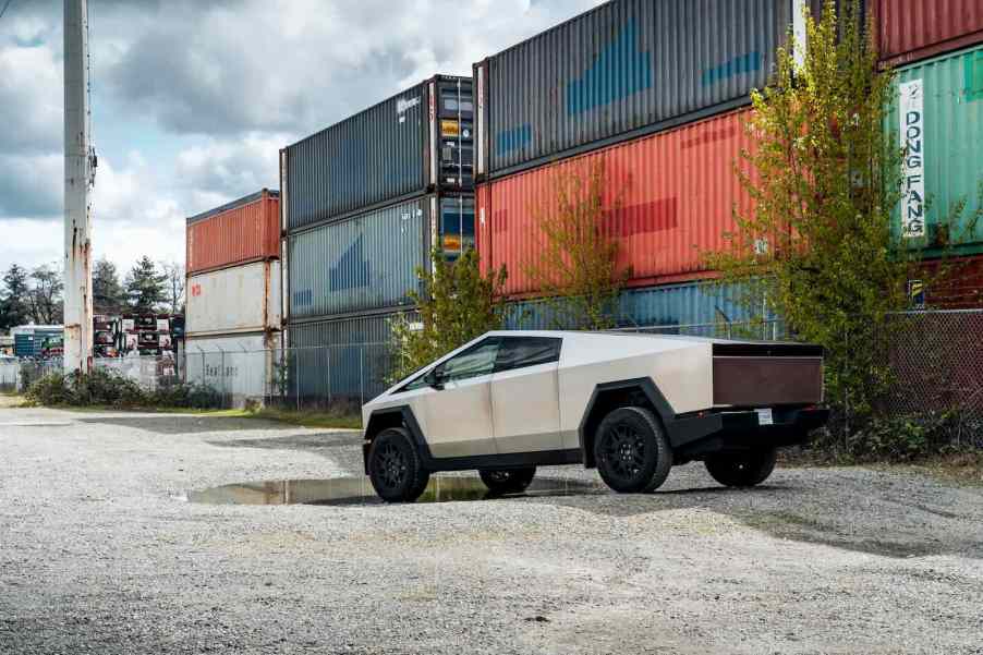 Tesla Cybertruck on gravel lot, shipping containers in the background.