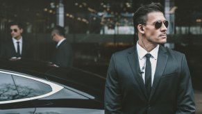 Bodyguard in a suit and tie wearing sunglasses standing outside a black vehicle