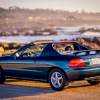 A dark-colored 1996 Honda Civic Del Sol parked on a rocky beach in left rear angle view with top off