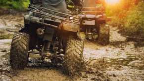 Two ATV riders crossing a dirt road, the setting sun in the background.